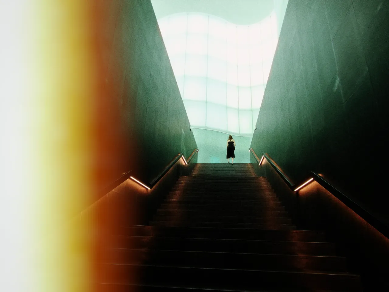 A lone figure at the top of a long dark stairway, under a tall window of white light, with left-edge burn at 50%