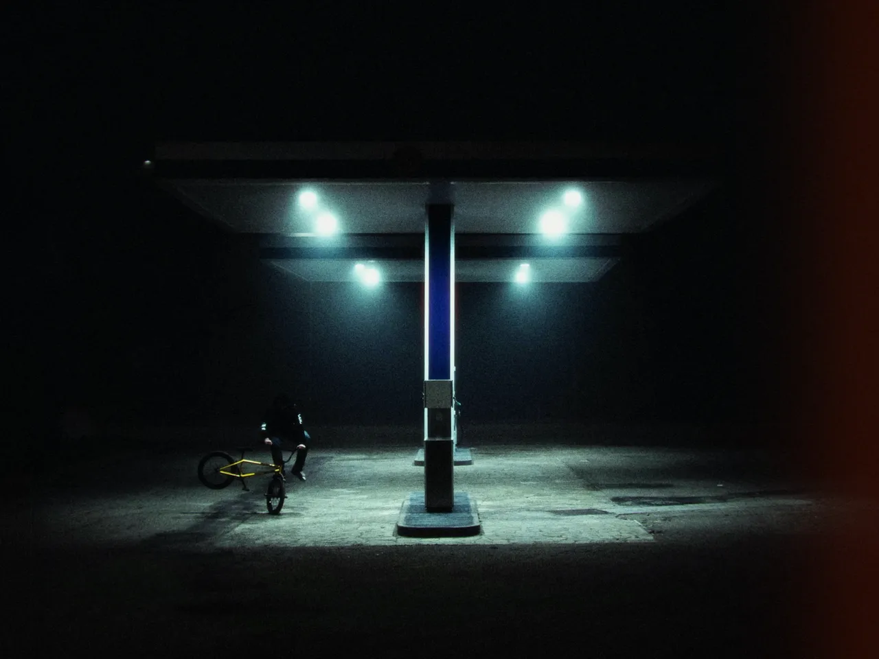 A rider on a bike under an empty gas station canopy at night, lit by cold blue lamps, with right-edge leak at 50%