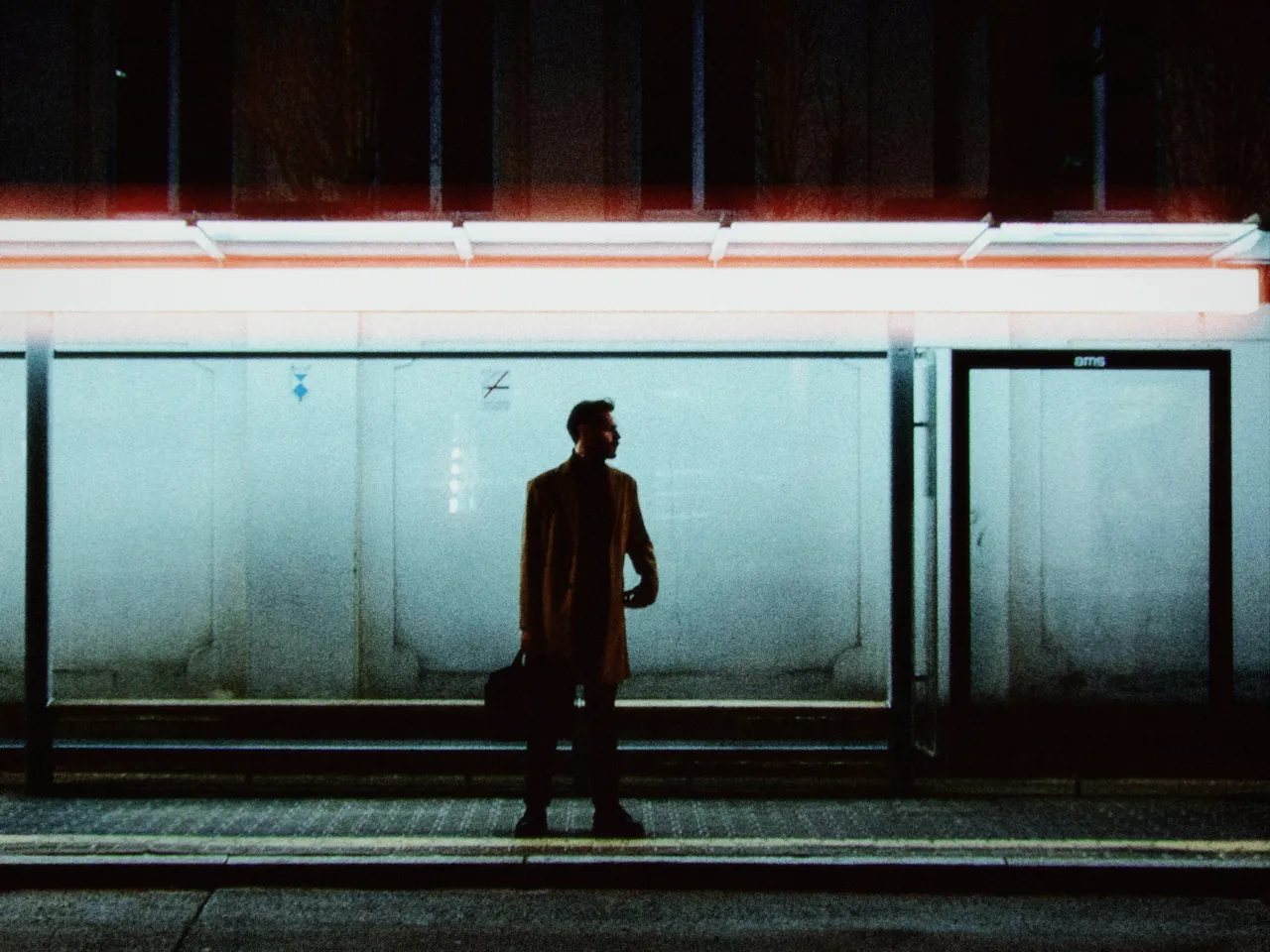 A man in a long coat waiting at a lit bus shelter at night, with push/pull at 0 stops